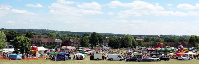 Panoramic view of large crowd of people enjoying large inflatables and stalls on Jessel Green, with distant views of countryside in the background